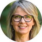Smiling woman with shoulder-length gray-blonde hair and rectangular glasses outdoors, wearing hoop earrings.